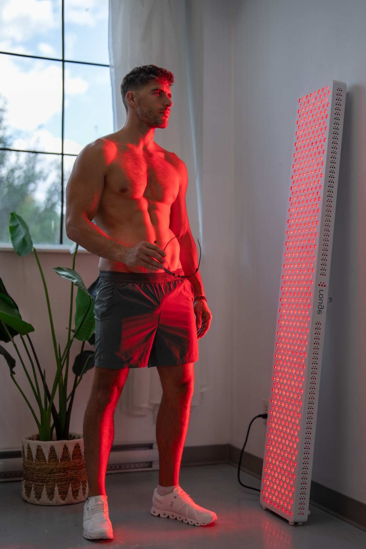 Shirtless man in shorts stands by a lit LED panel in a bright room, bathed in red light.