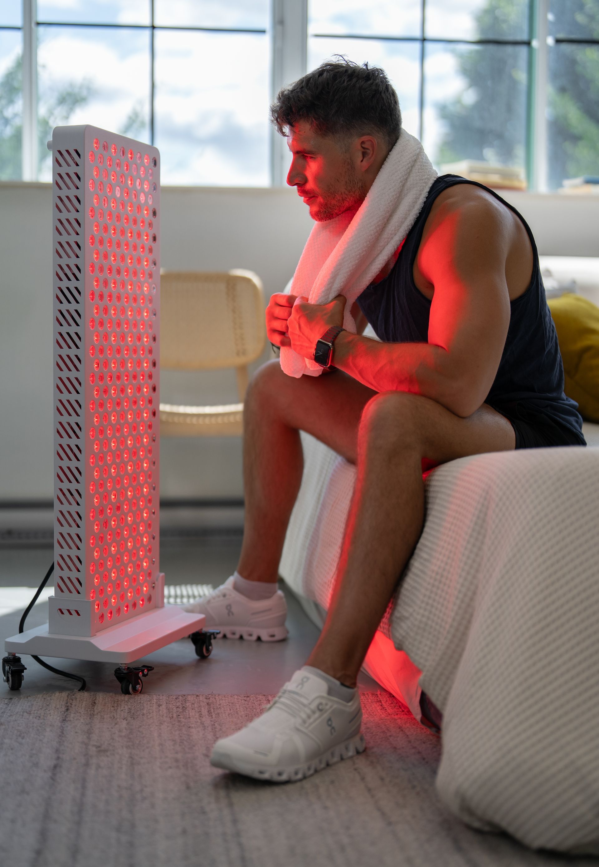 Person sitting beside a glowing red tower heater in a bright bedroom, wrapped in a towel and holding a mug.