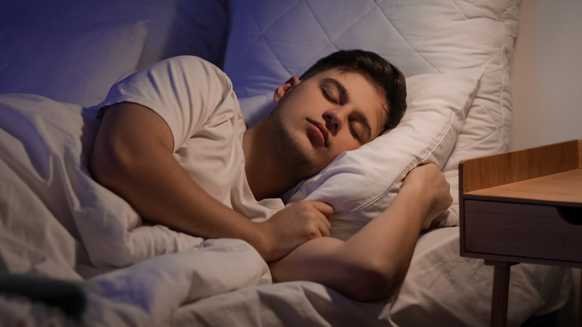 Person sleeping in bed beside a wooden nightstand, head on a white pillow under white sheets.