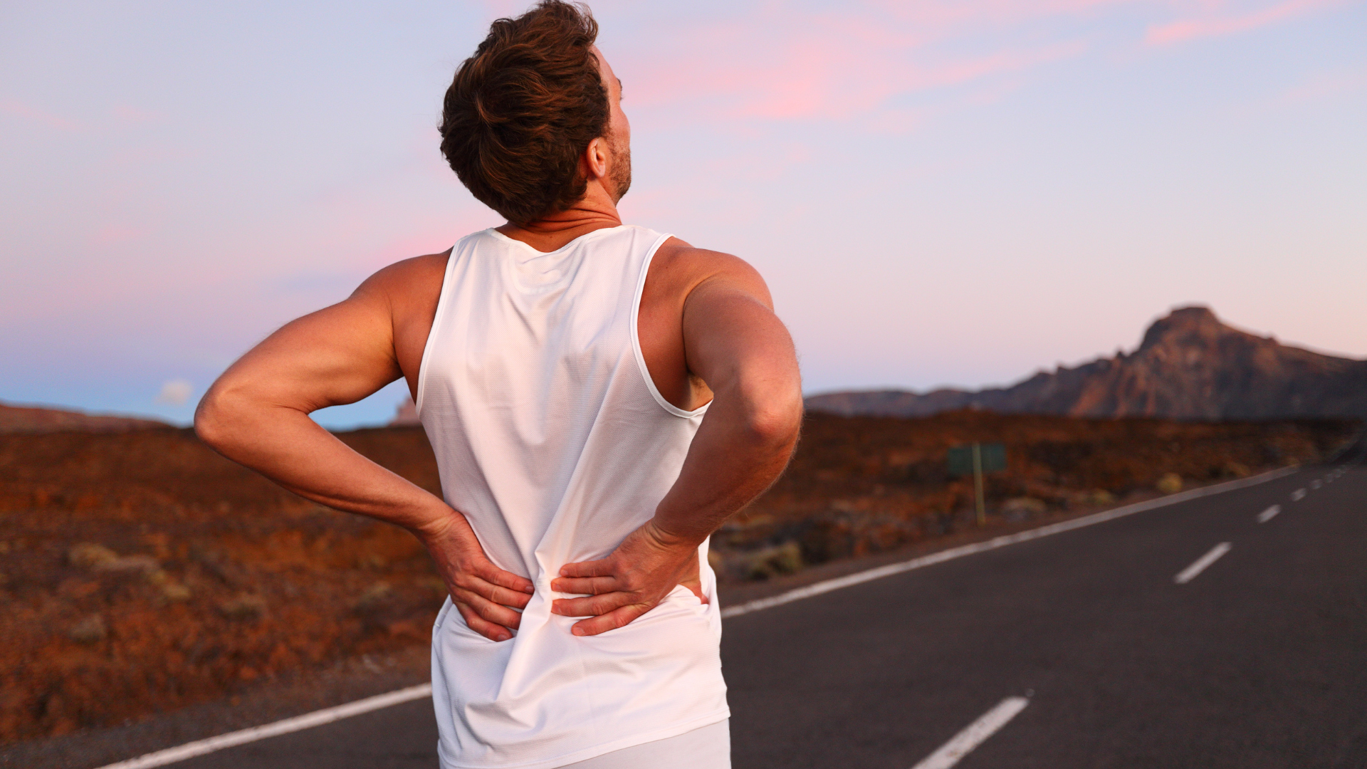 Person standing on a road at sunset, holding their lower back and looking toward distant hills.