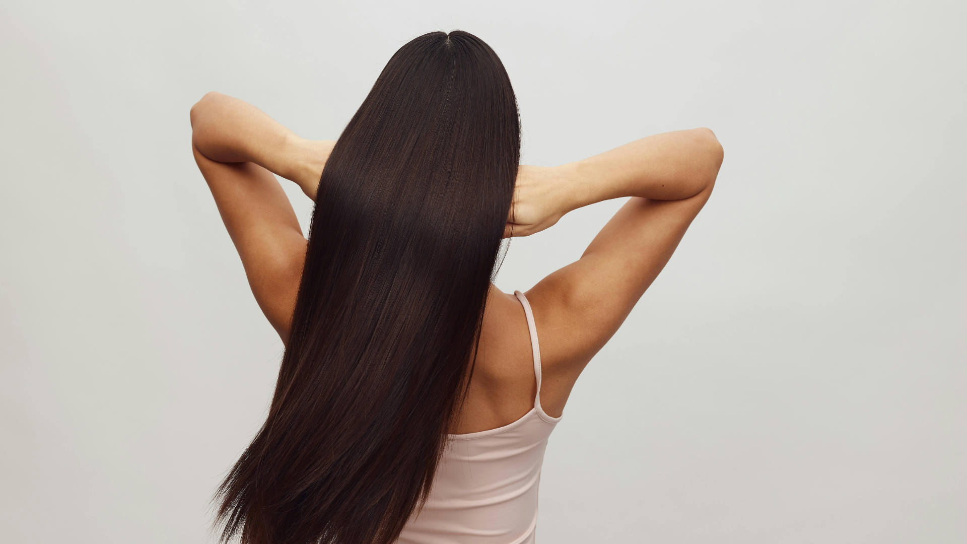 Woman with long dark hair standing with hands behind her head against a plain white background