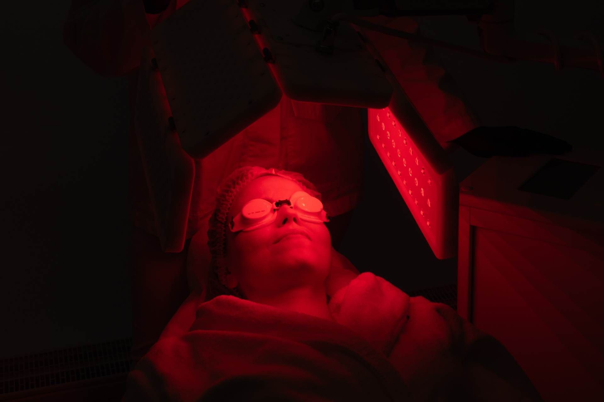 Person lit by red light, wearing glasses, lying under a dark machine or device.