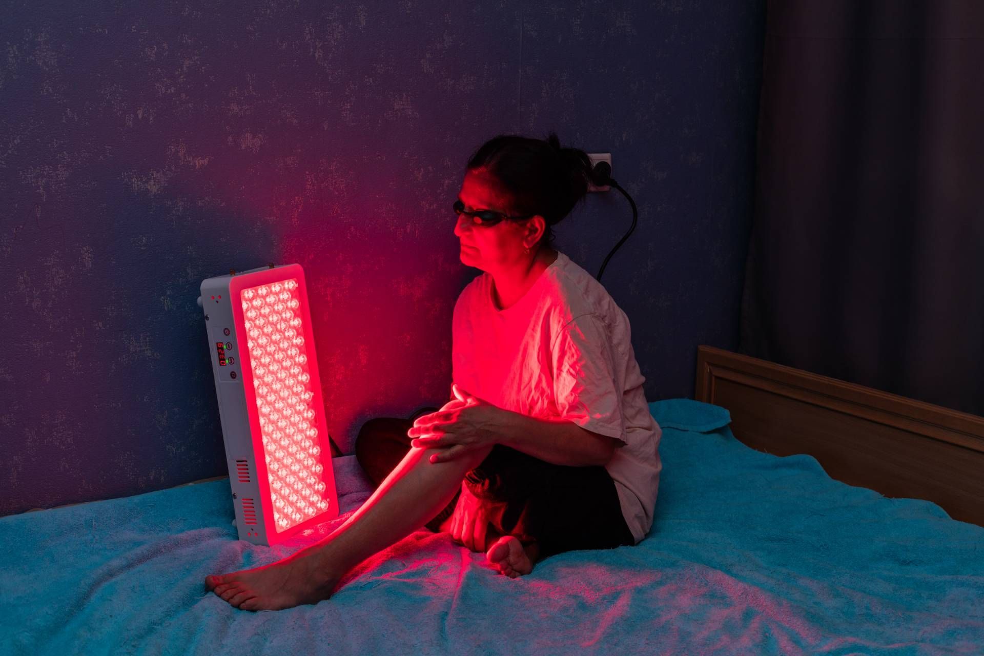 Person sitting beside a glowing red light therapy panel on a bed in a dark room