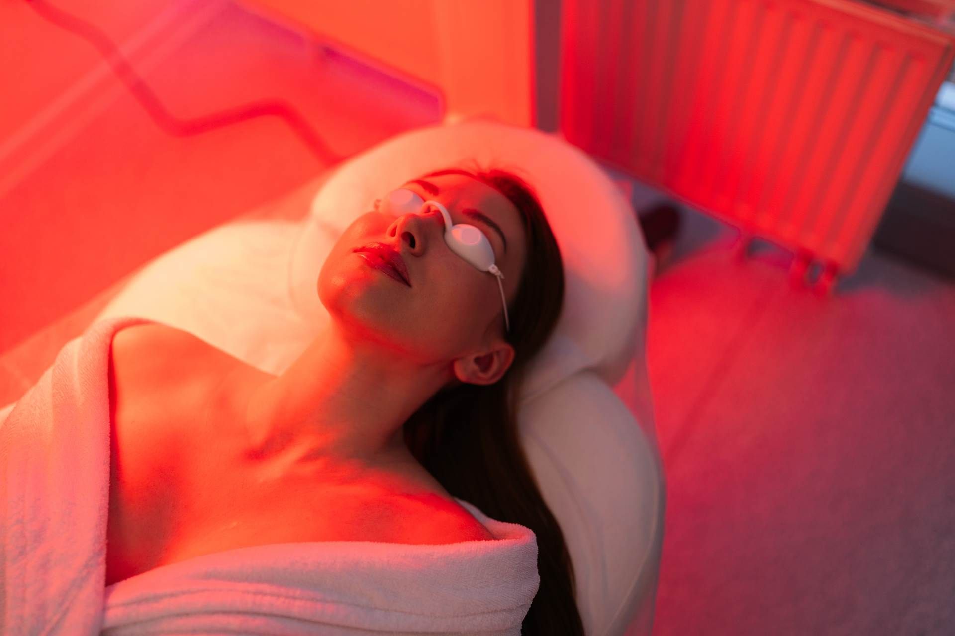 Person lying on a white massage bed under red light therapy, wearing protective goggles.