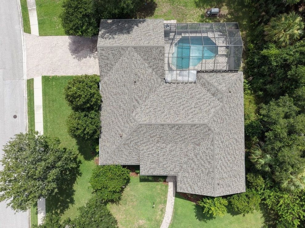 Aerial view of a house roof with gray shingles and dark blue sections, surrounded by trees and greenery.