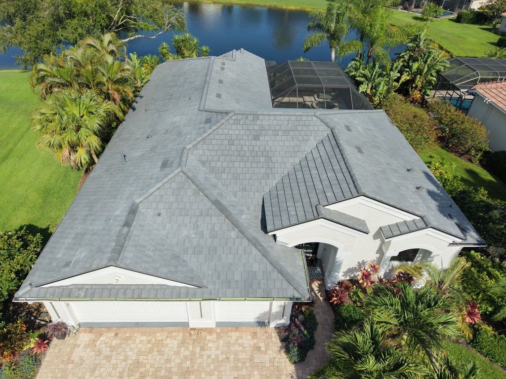 Aerial view of a gray-roofed house beside a lake, surrounded by tropical landscaping.