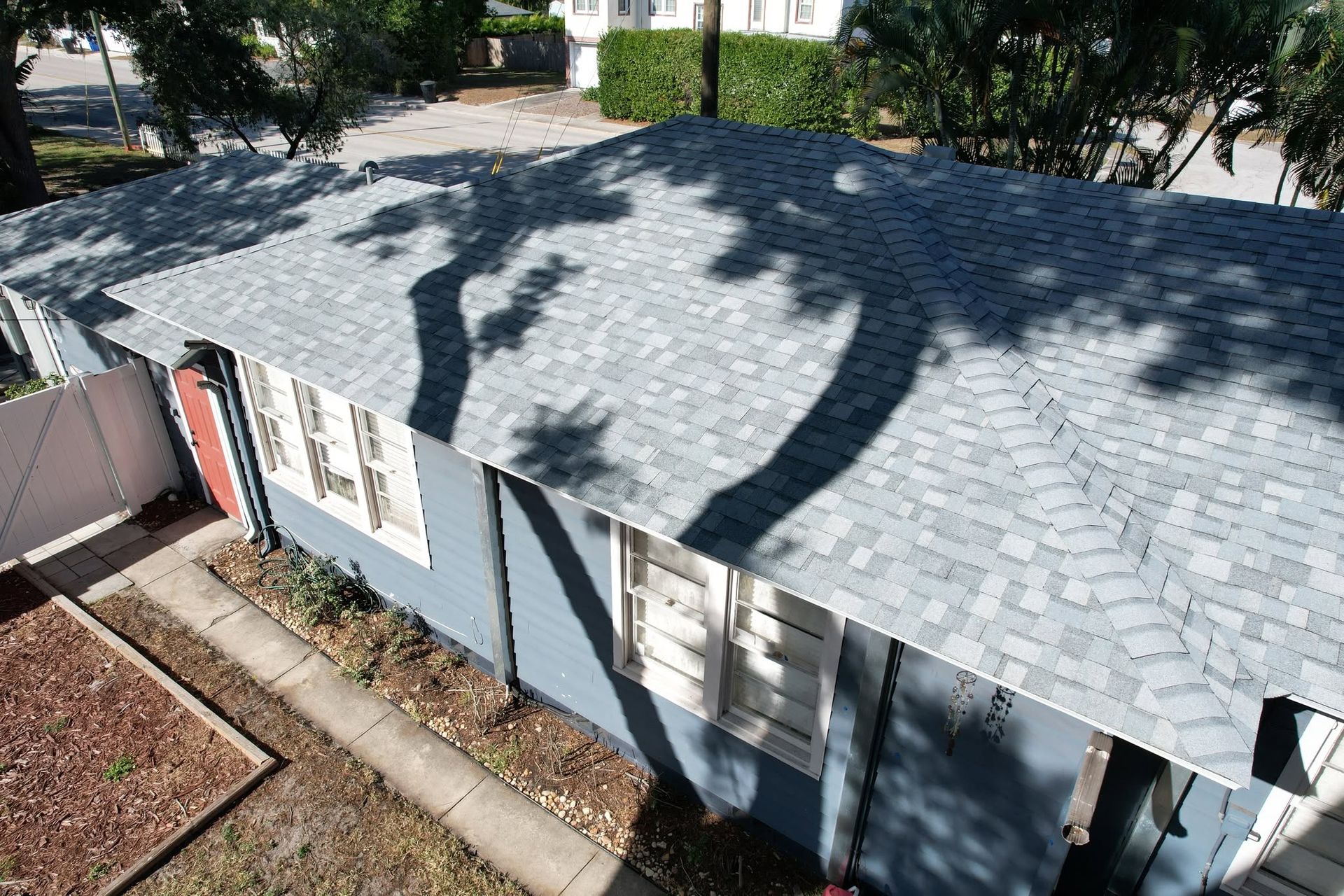 Roof of a light blue house with gray shingles and tree shadows, viewed from above