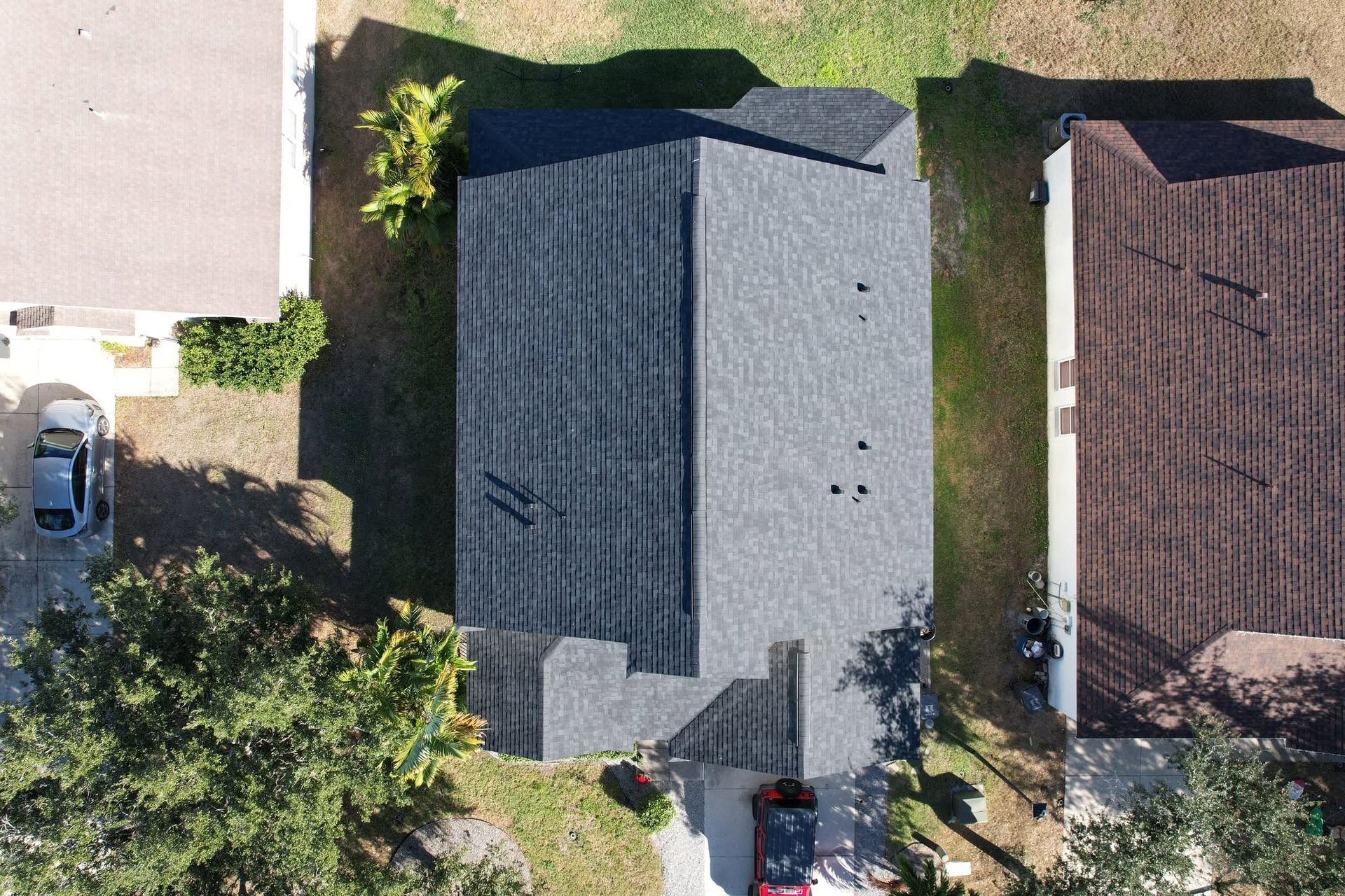 Aerial view of two houses with dark roofs, driveways, parked cars, and trees along a residential street