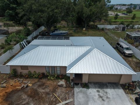 Aerial view of a white-roofed house with a blue section and surrounding trees and yard