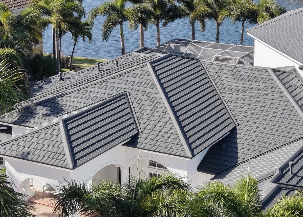 Aerial view of a gray tiled roof with palm trees and water in the background.