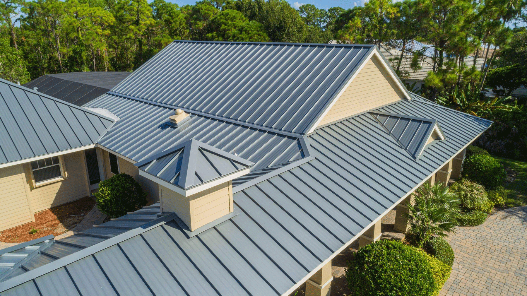 Aerial view of a house with a gray metal roof surrounded by green trees and landscaping