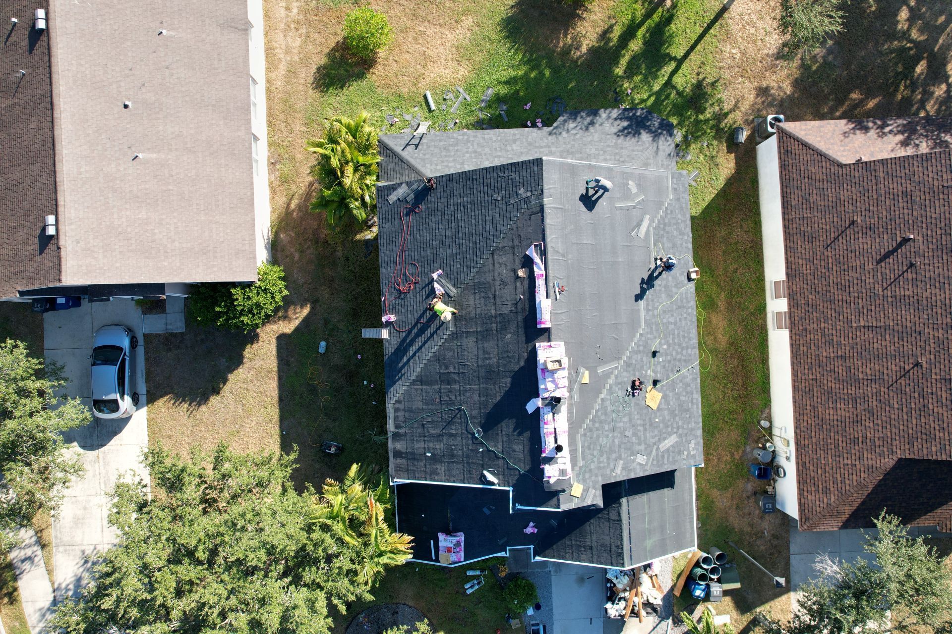 Aerial view of roofers working on a gray shingle roof beside neighboring houses and a driveway.