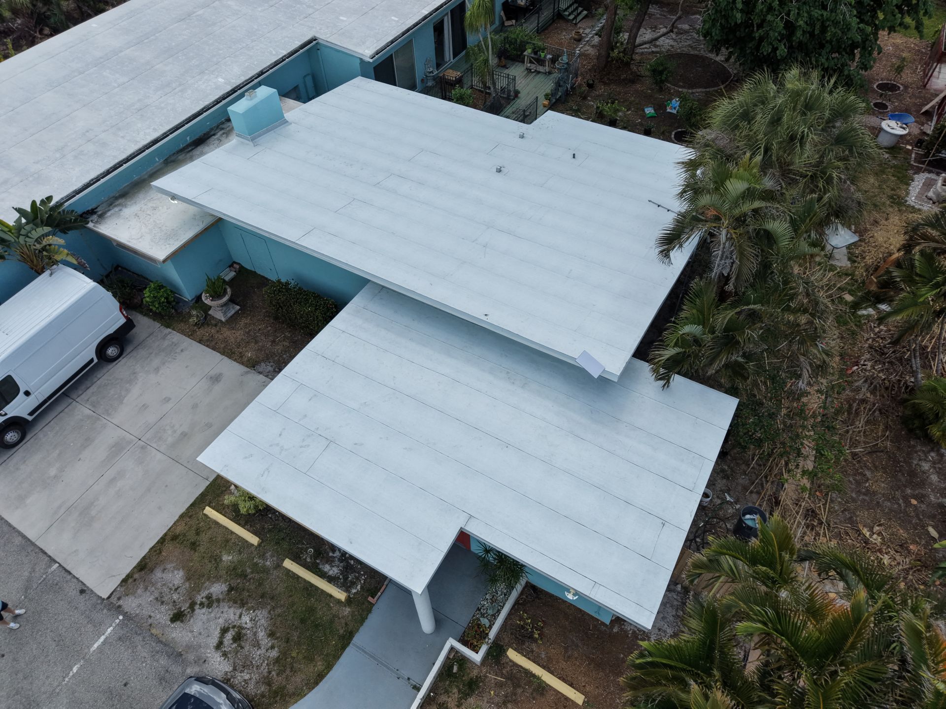 Aerial view of a white-roofed house with a carport, driveway, and surrounding trees and shrubs.