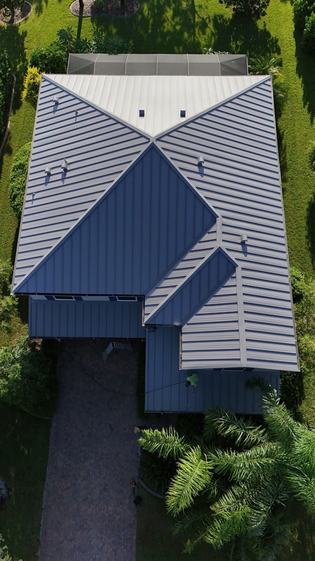 Aerial view of a house with a gray tile roof and dark blue metal roof section, surrounded by trees.