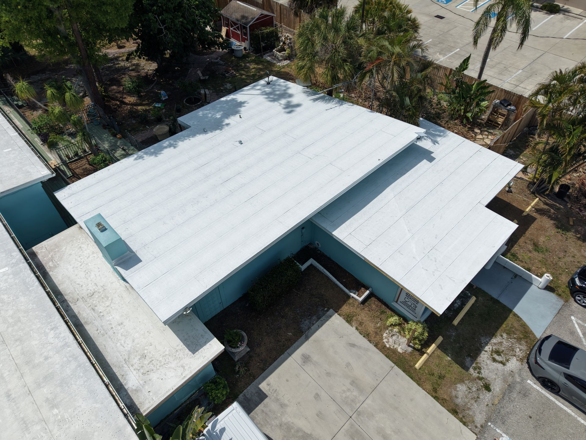 Aerial view of a single-story house with a white roof and driveway, surrounded by trees and yard.