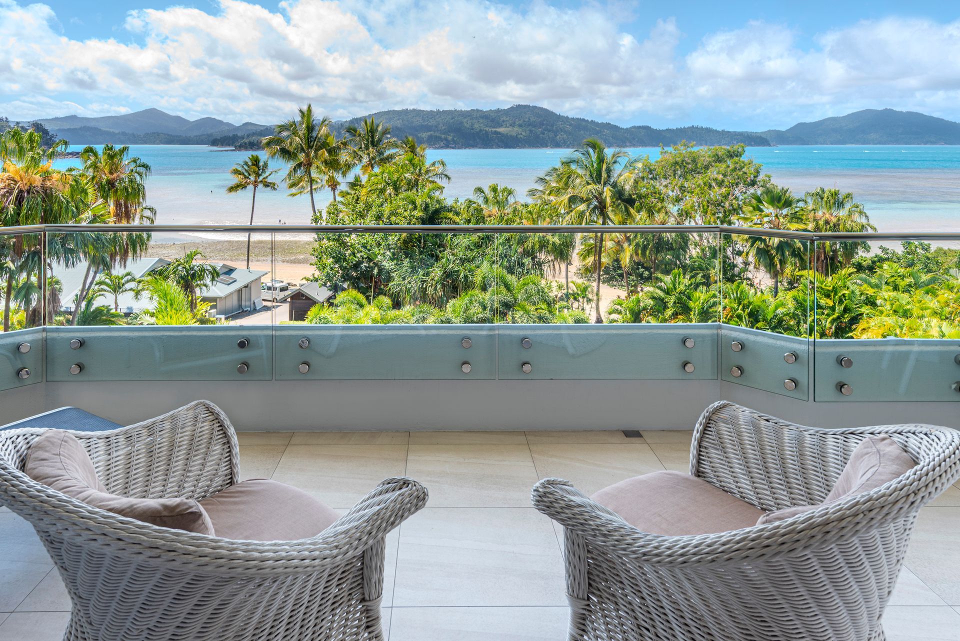 Balcony with wicker chairs overlooking a tropical beach, palm trees, and blue ocean mountains