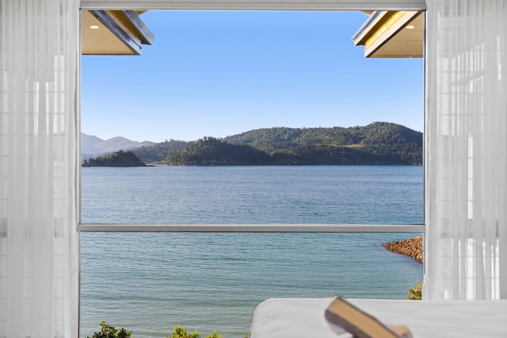 Balcony view of a calm blue lake with distant hills, framed by white curtains and a clear sky