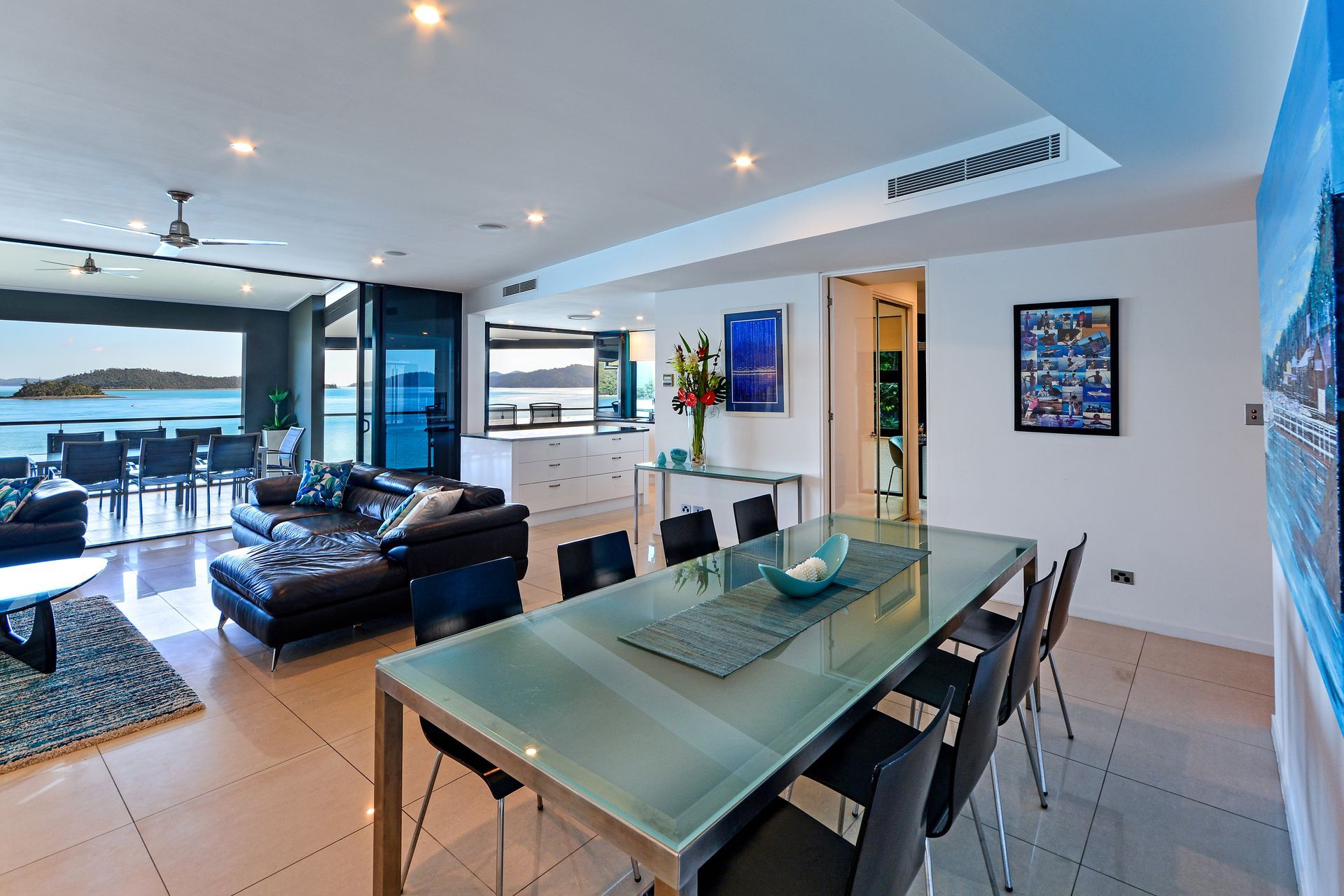 Modern open-plan living room and dining area with black leather sofa, glass table, and ocean view