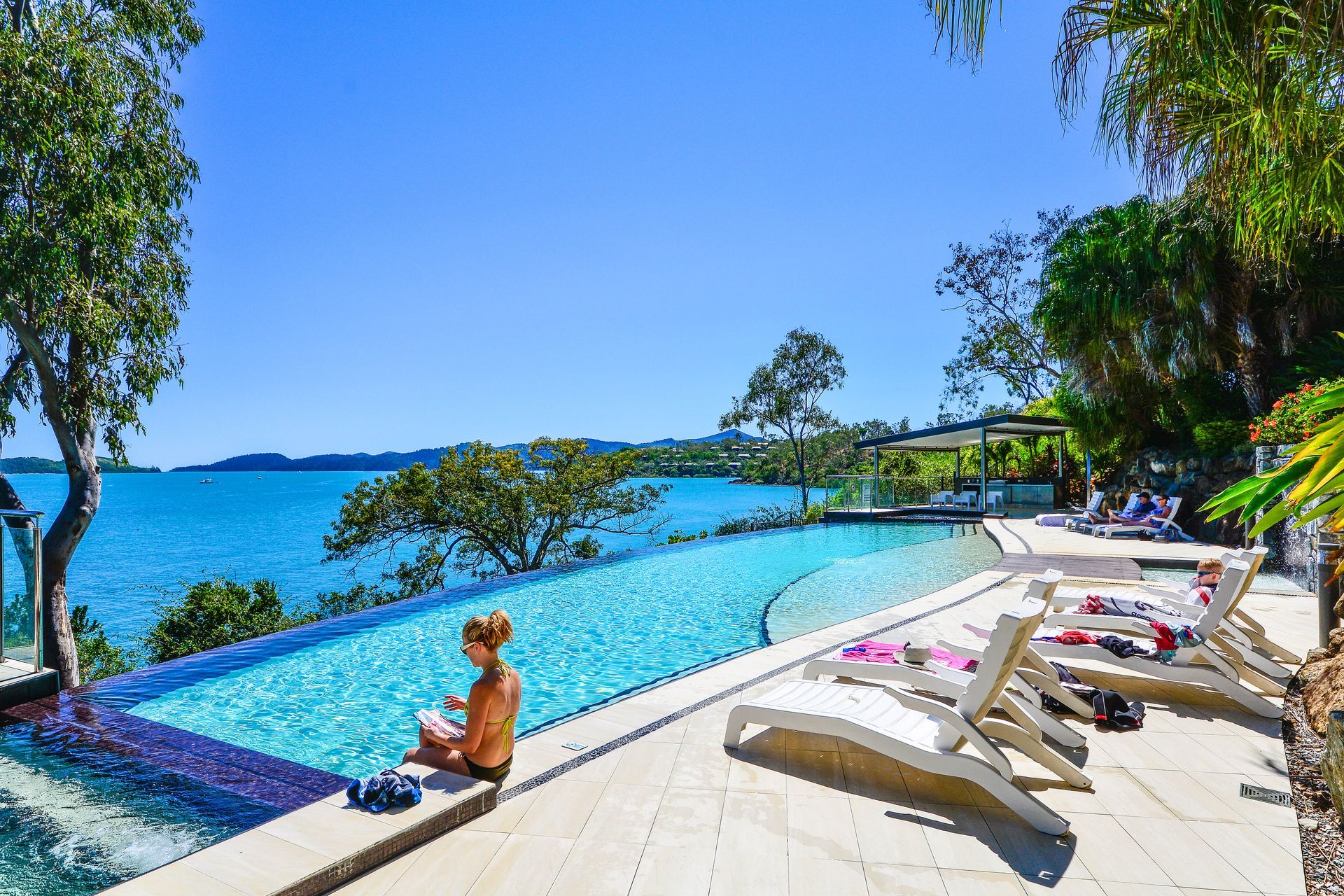 Infinity pool overlooking the ocean with lounge chairs and a person relaxing by the water