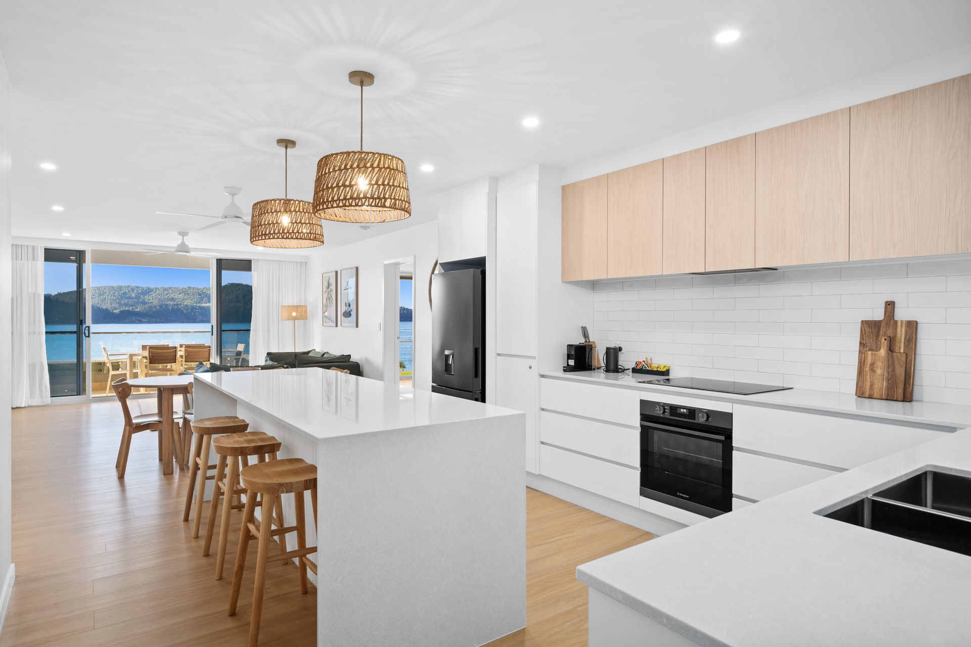Bright modern kitchen with white island, wood cabinets, pendant lights, and ocean view beyond dining area