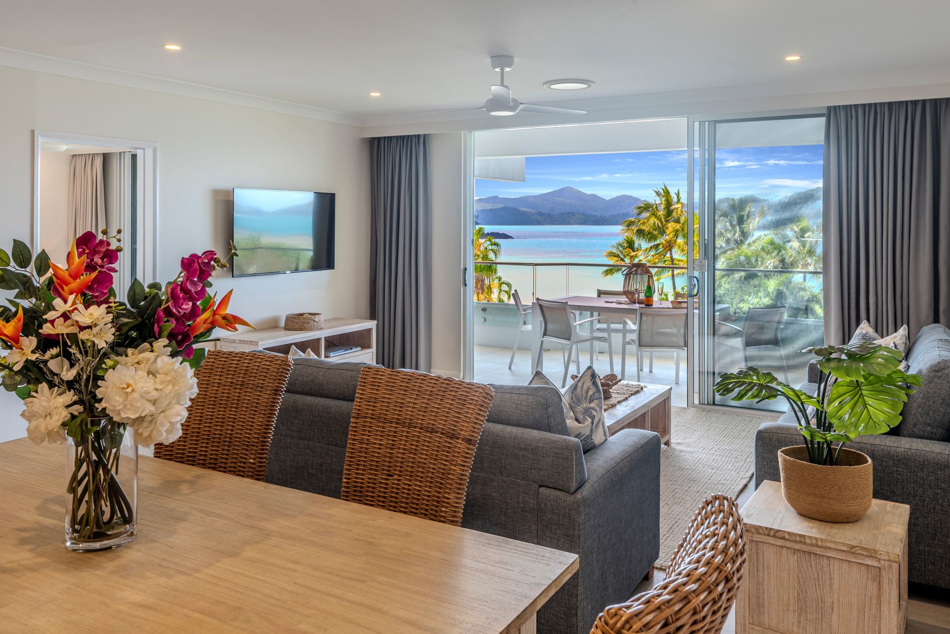 Bright living room with ocean-view balcony, gray sofa, dining table, and potted plants