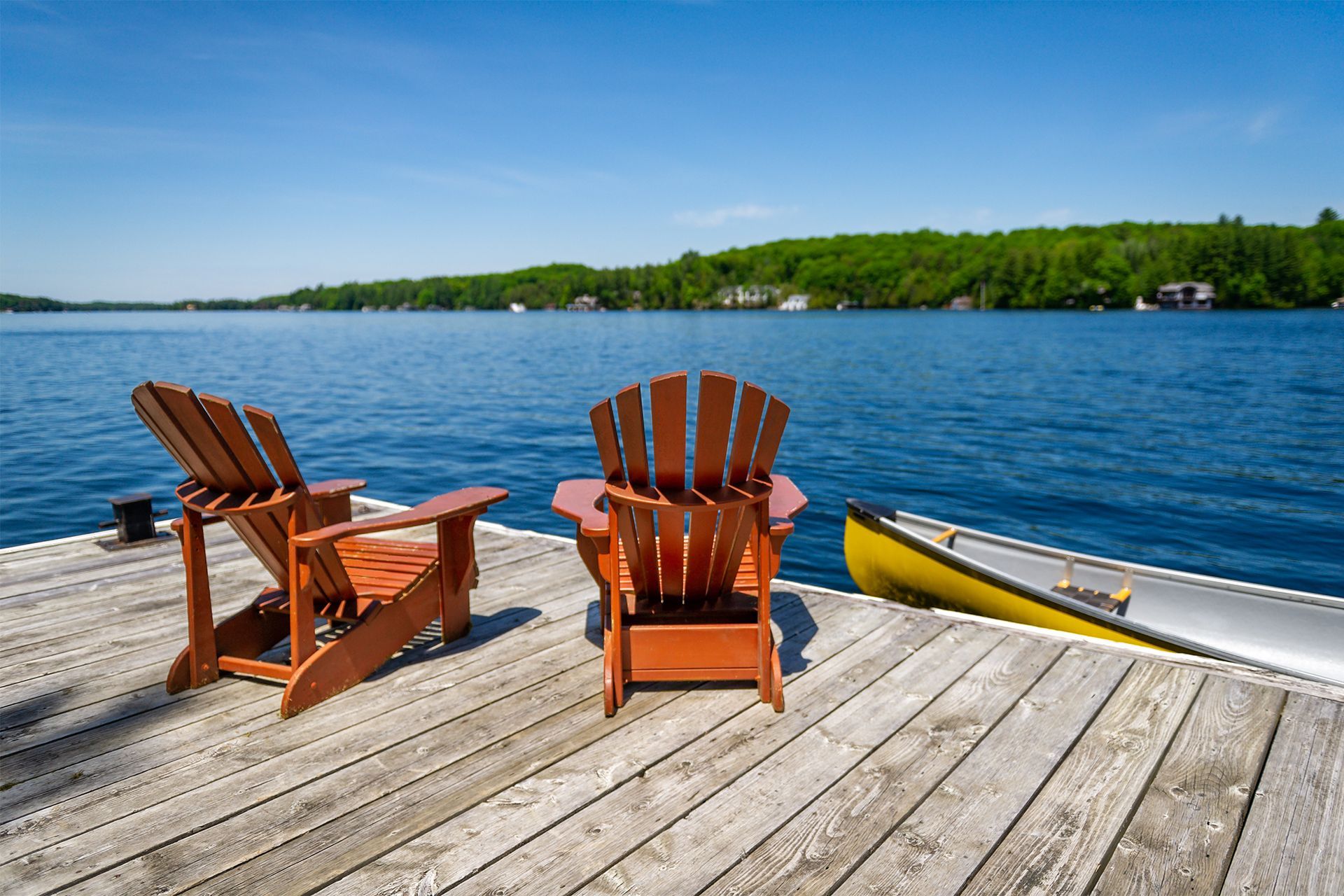 Two wooden Adirondack chairs face a lake from a dock, canoe at the side, blue sky, sunny day.