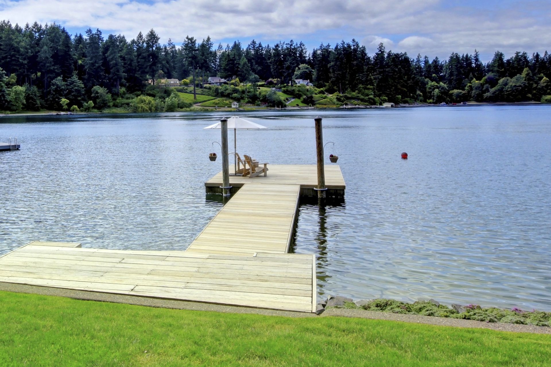 Wooden dock with chairs and umbrella on a lake, surrounded by trees and blue sky.