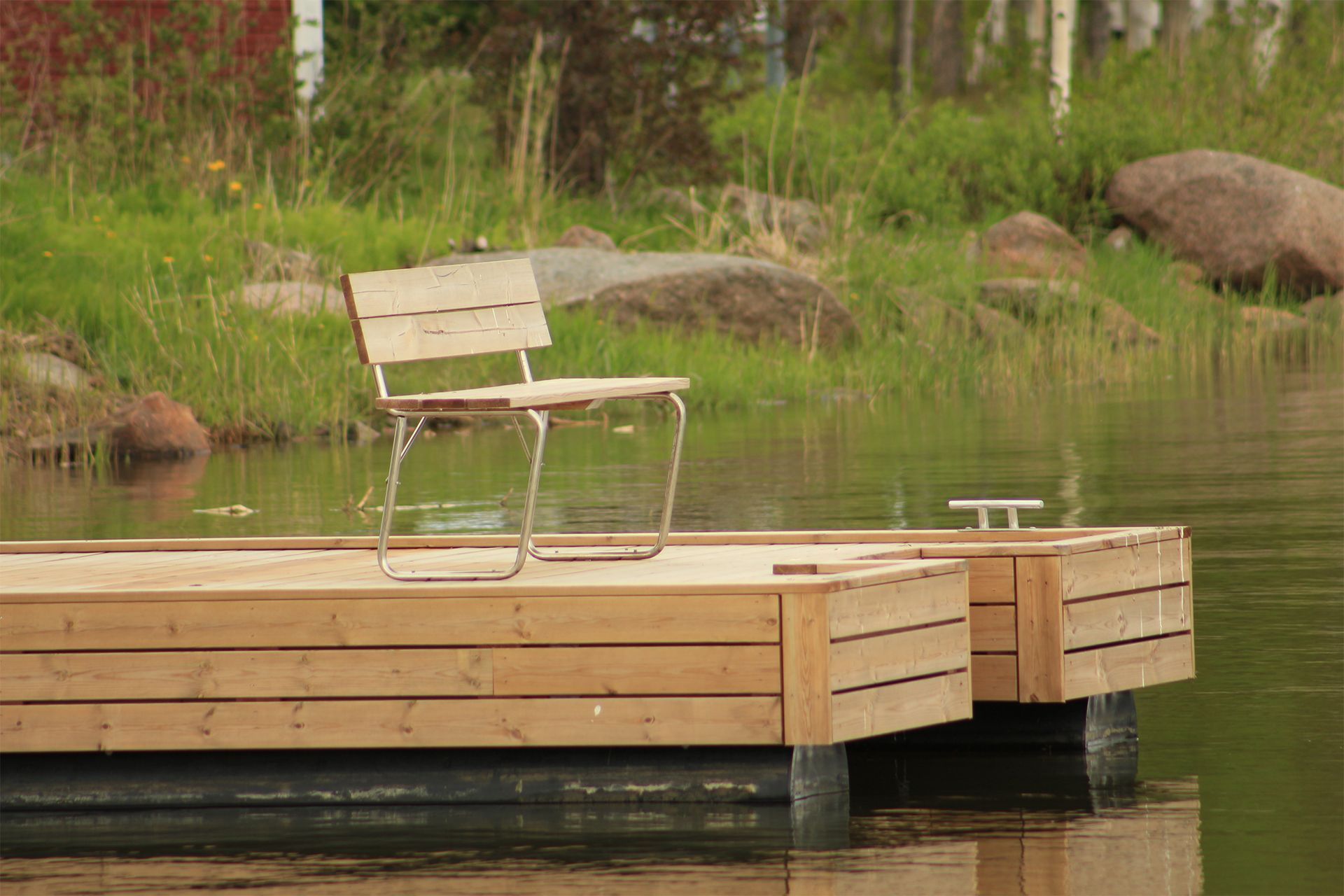 Wooden chair on a wooden dock over calm water. Green grass and rocks in background.