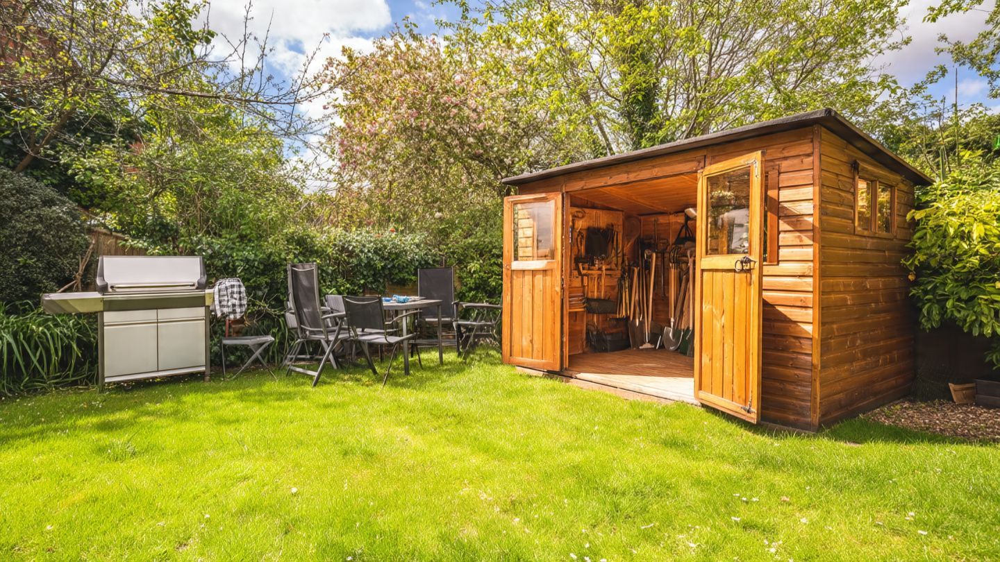 Wooden garden shed with open doors beside outdoor seating on a grassy lawn, surrounded by trees