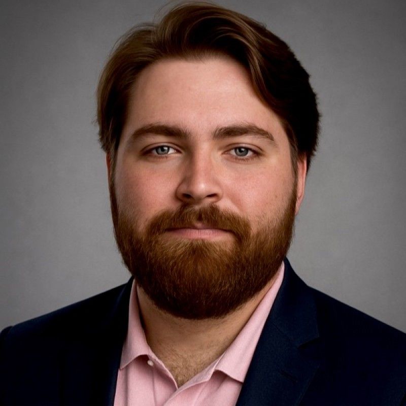 Studio headshot of a person with brown hair and beard, wearing a dark blazer and pink shirt on a gray background