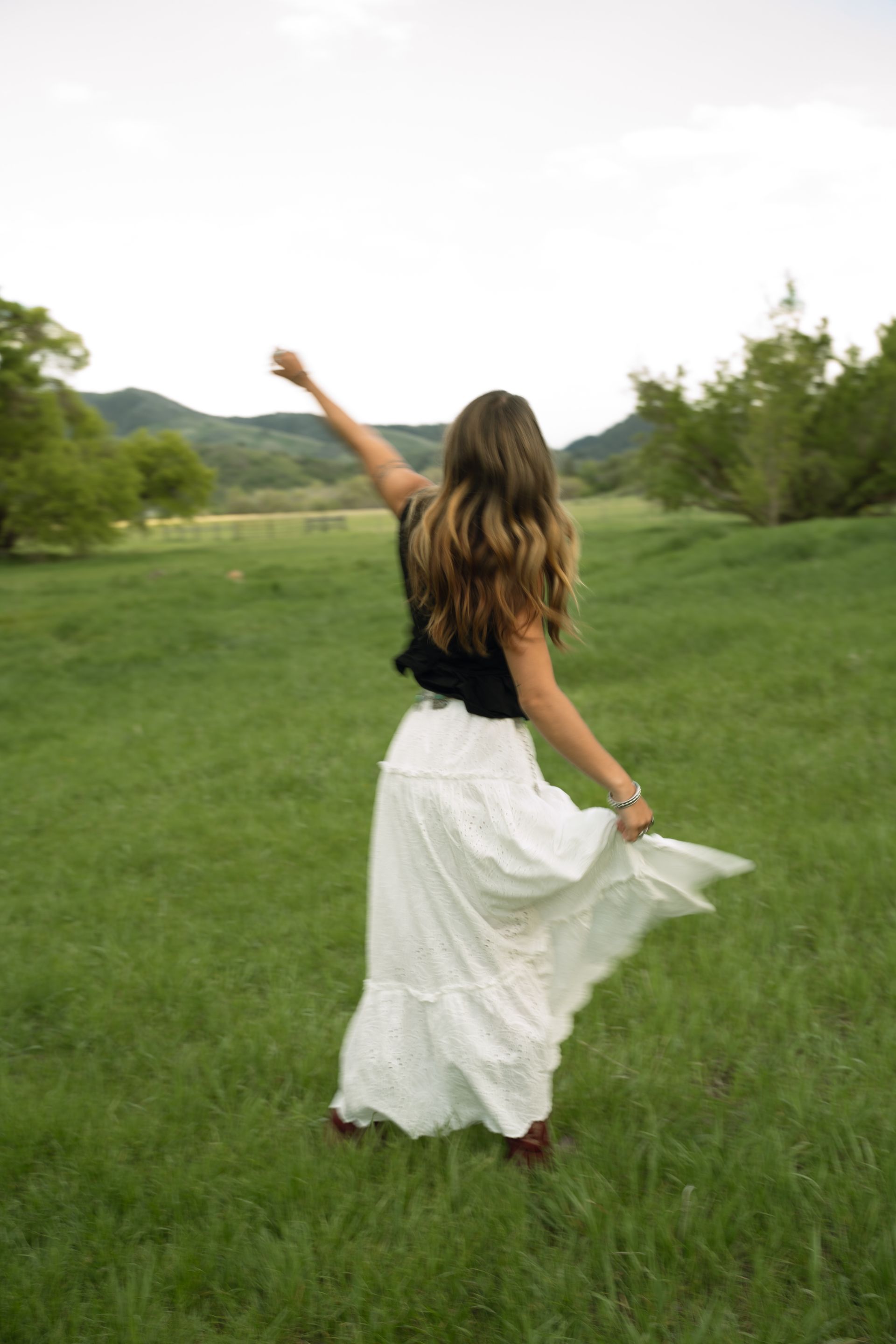 A woman in a white skirt is standing in a grassy field with her arms outstretched.