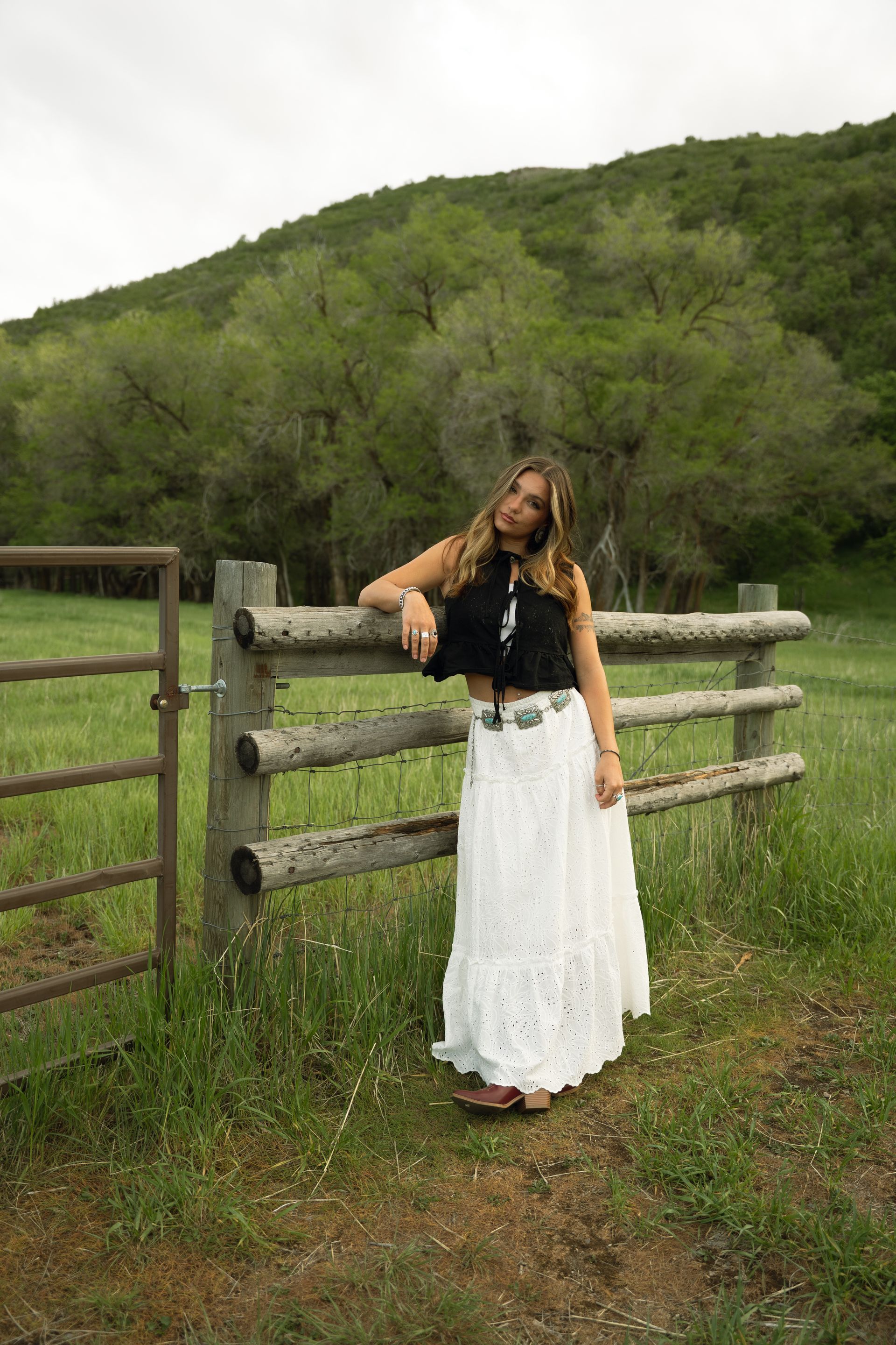 A woman in a white skirt is leaning against a wooden fence in a field.