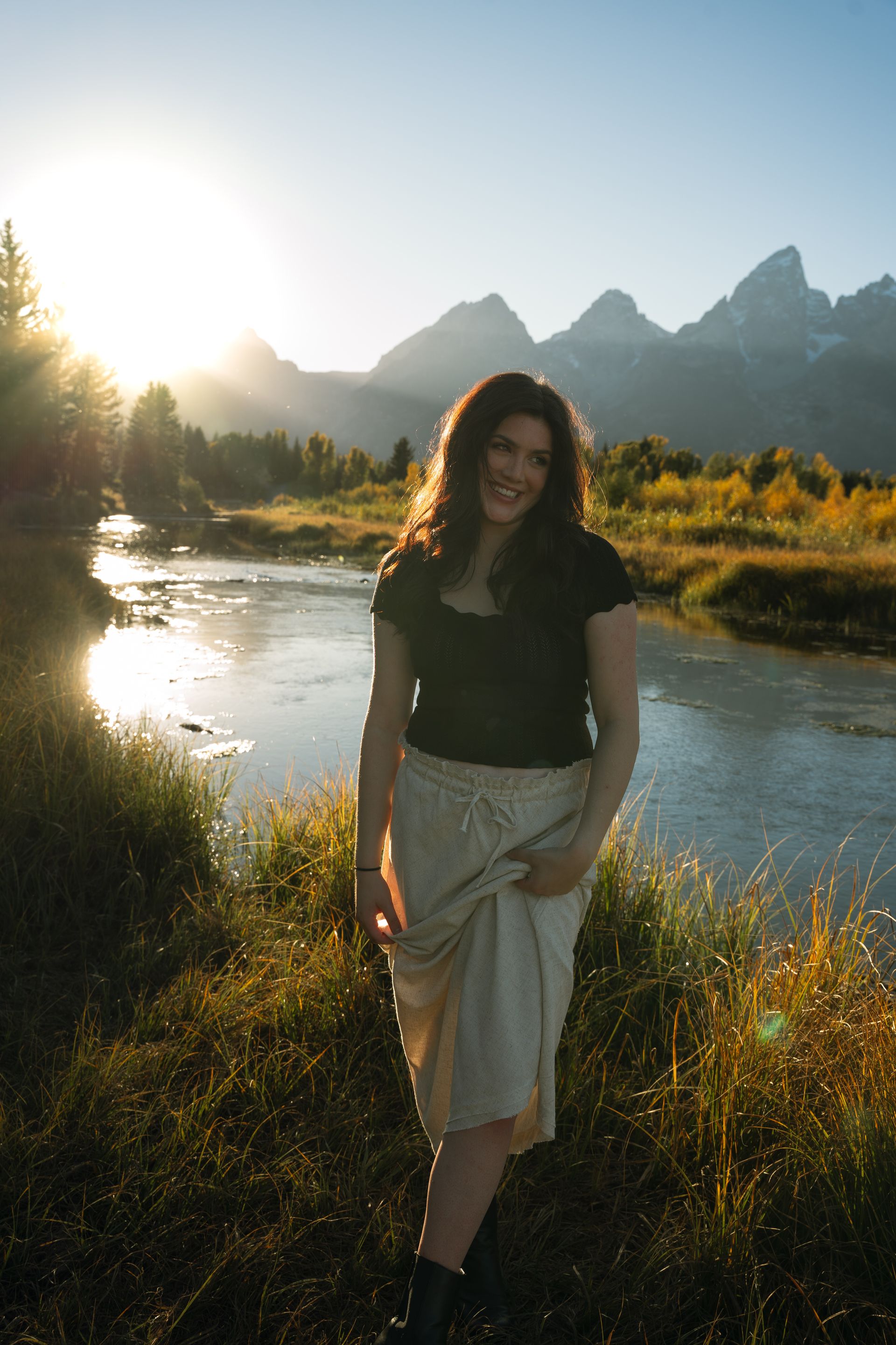 A woman is standing in front of a river with mountains in the background.