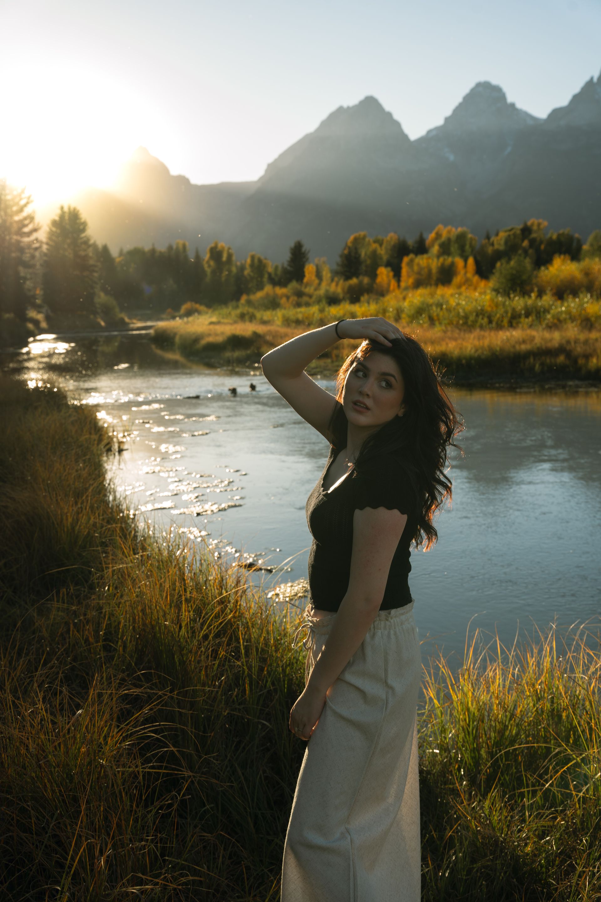 A woman is standing next to a river with mountains in the background.