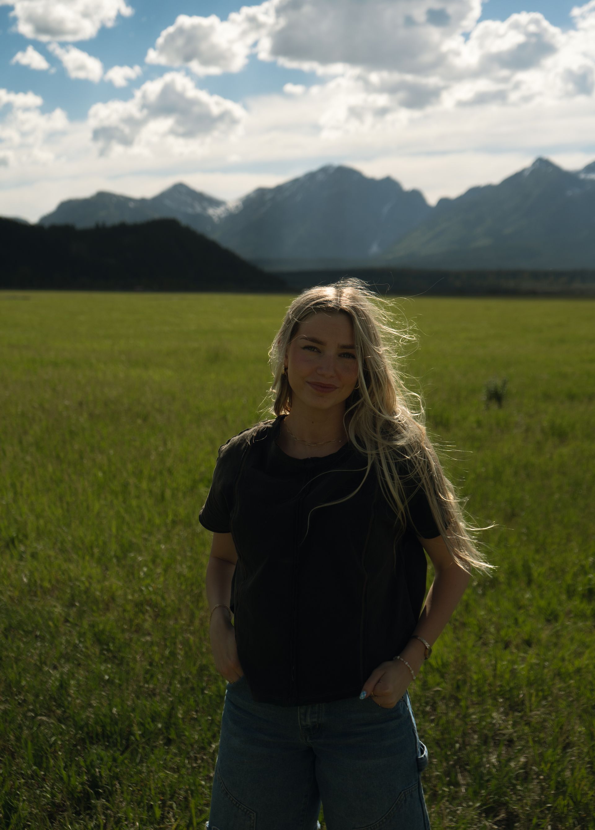 A woman is standing in a grassy field with mountains in the background.
