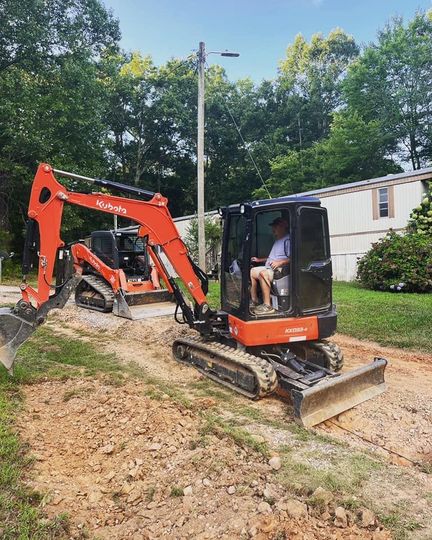 Orange mini excavator on a dirt path beside a house, with an operator inside and a bucket blade raised.