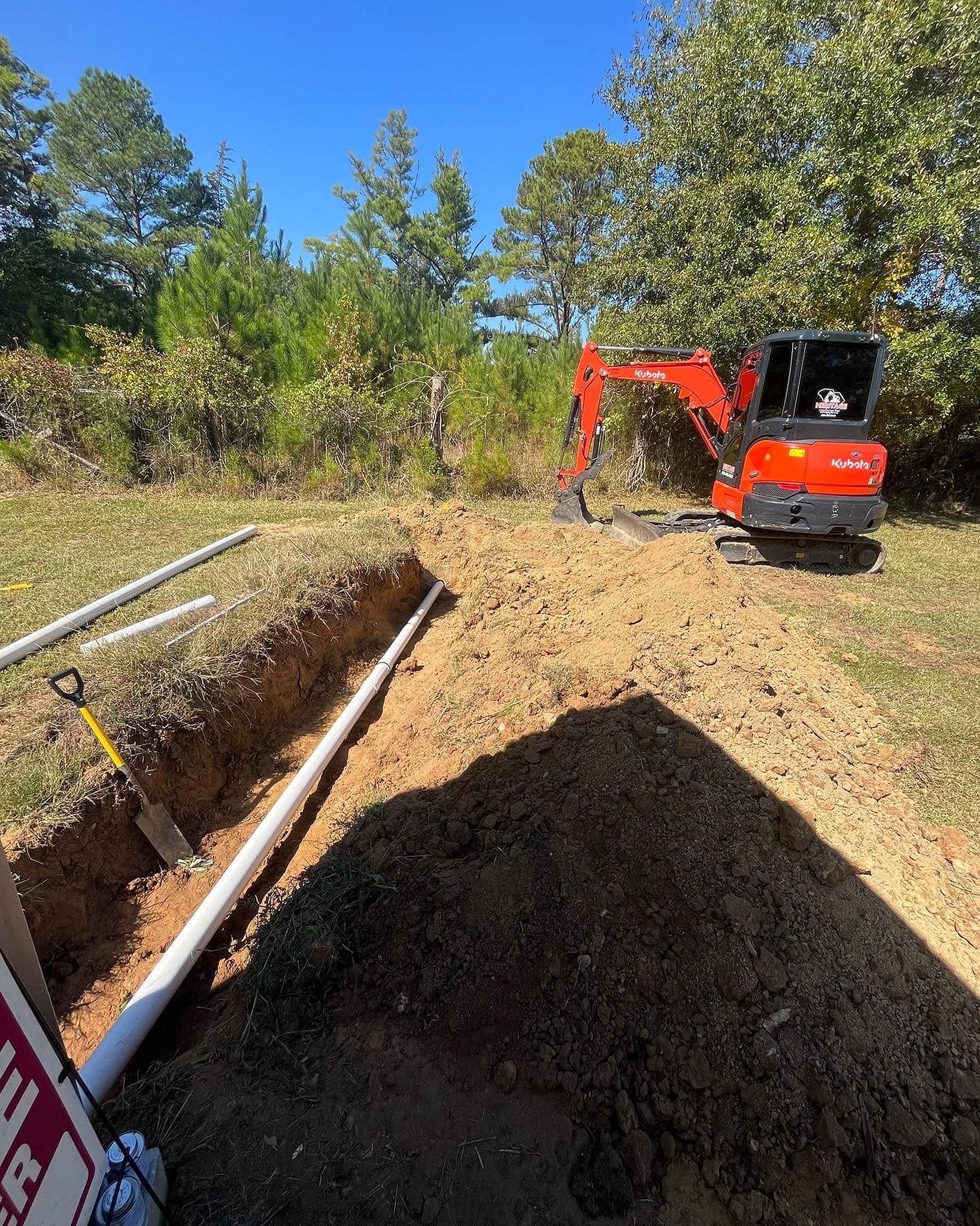 Orange excavator beside a trench and dirt pile on a wooded construction site