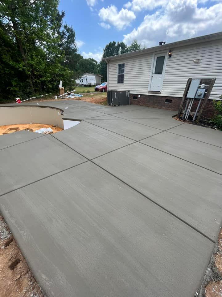 Freshly poured concrete patio beside a white house, with trees and driveway in the background