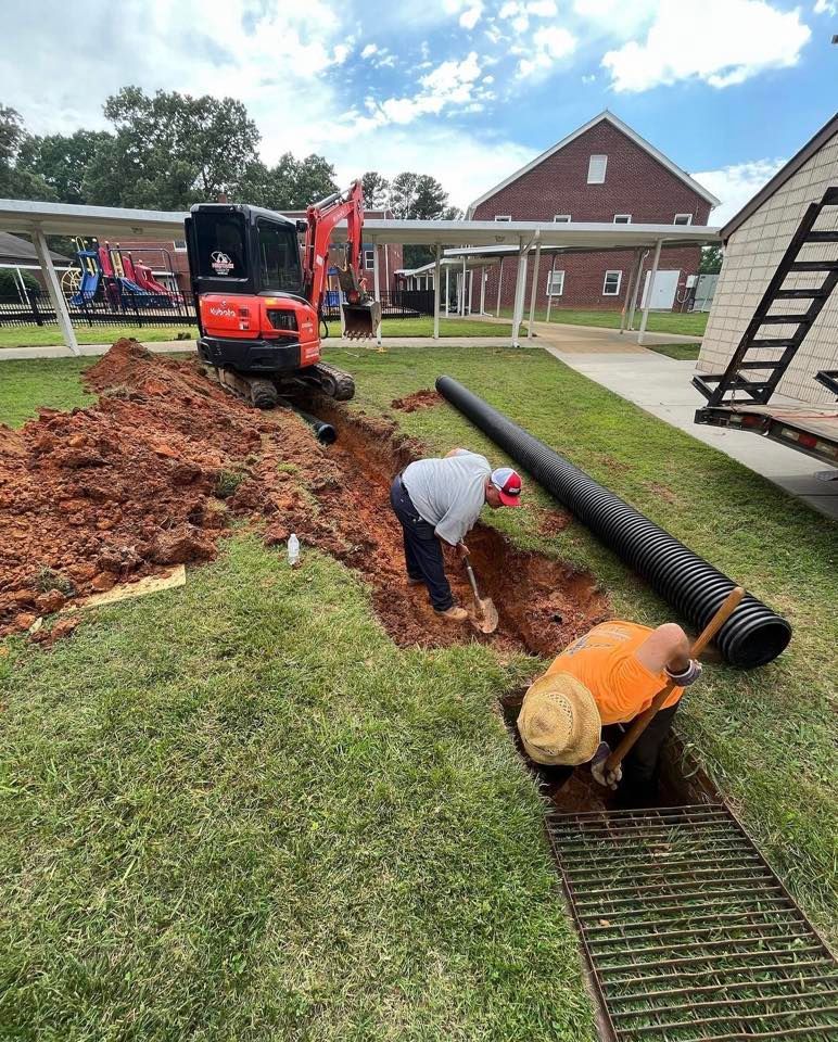 Workers dig a trench beside a red mini excavator, laying black pipe in a grassy yard near houses.