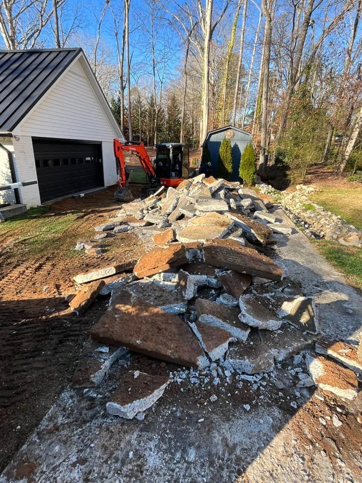 Demolition debris piled along a driveway beside a garage, with an excavator and trees in the background