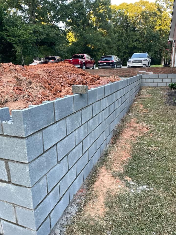 Gray cinder block retaining wall beside a dirt mound, with parked vehicles and trees in the background