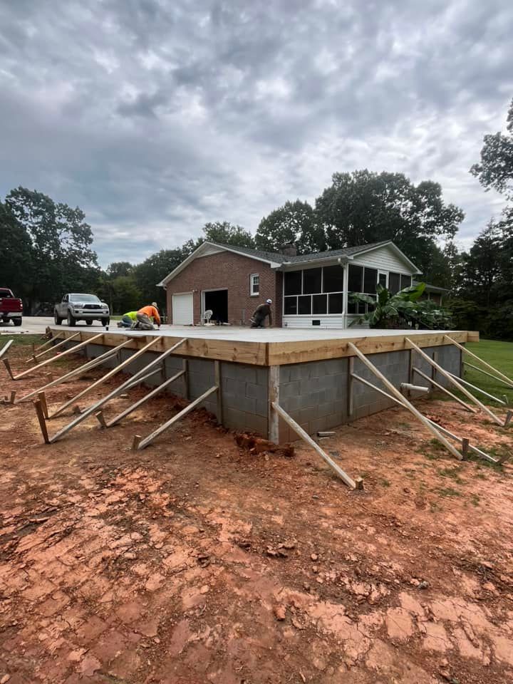 House foundation under construction with wooden braces on muddy red soil beneath a cloudy sky