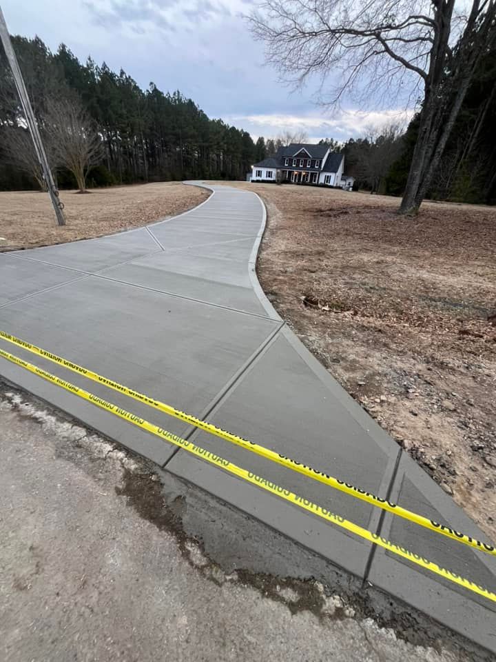 New concrete sidewalk curves through a grassy roadside area toward houses, with yellow tactile warning strips at the edge.