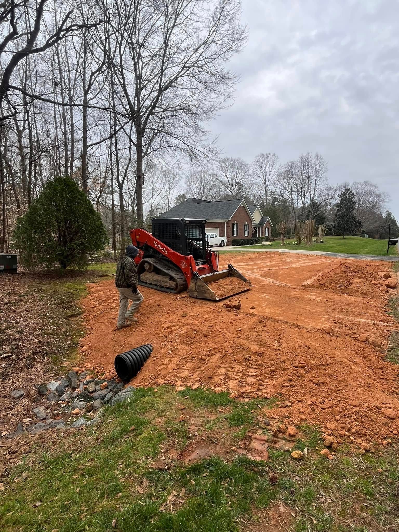 Red excavator grading a dirt driveway near a house in a wooded yard.
