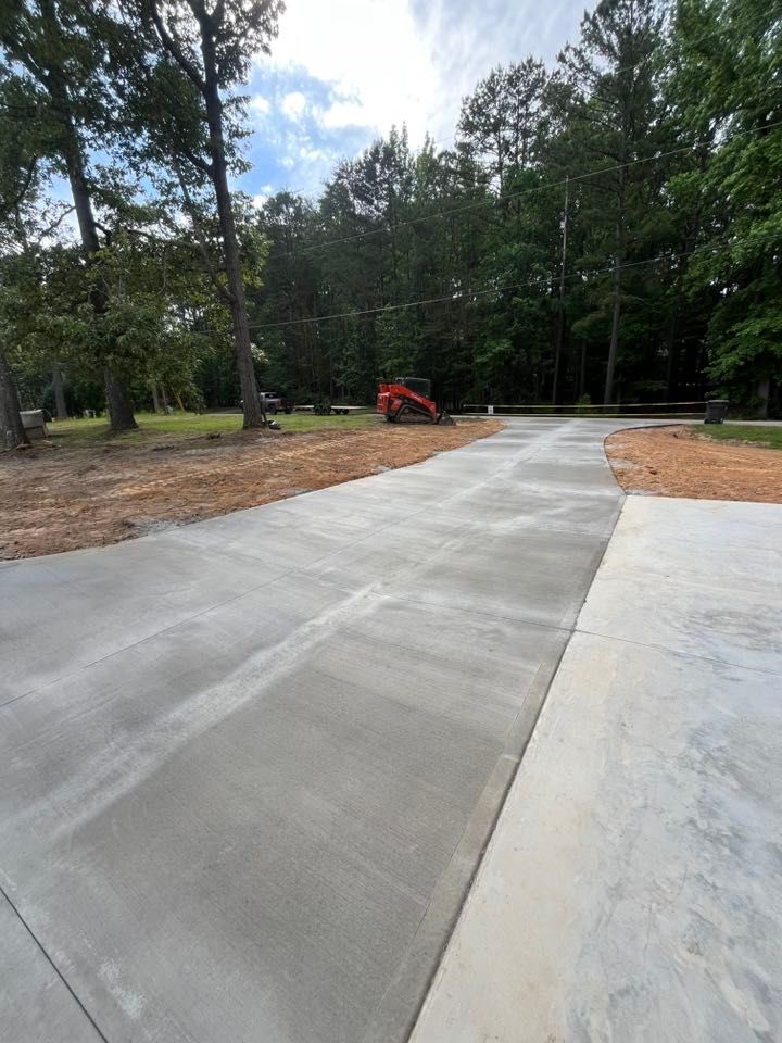 Fresh concrete driveway curving through a wooded area, with a small orange excavator in the distance
