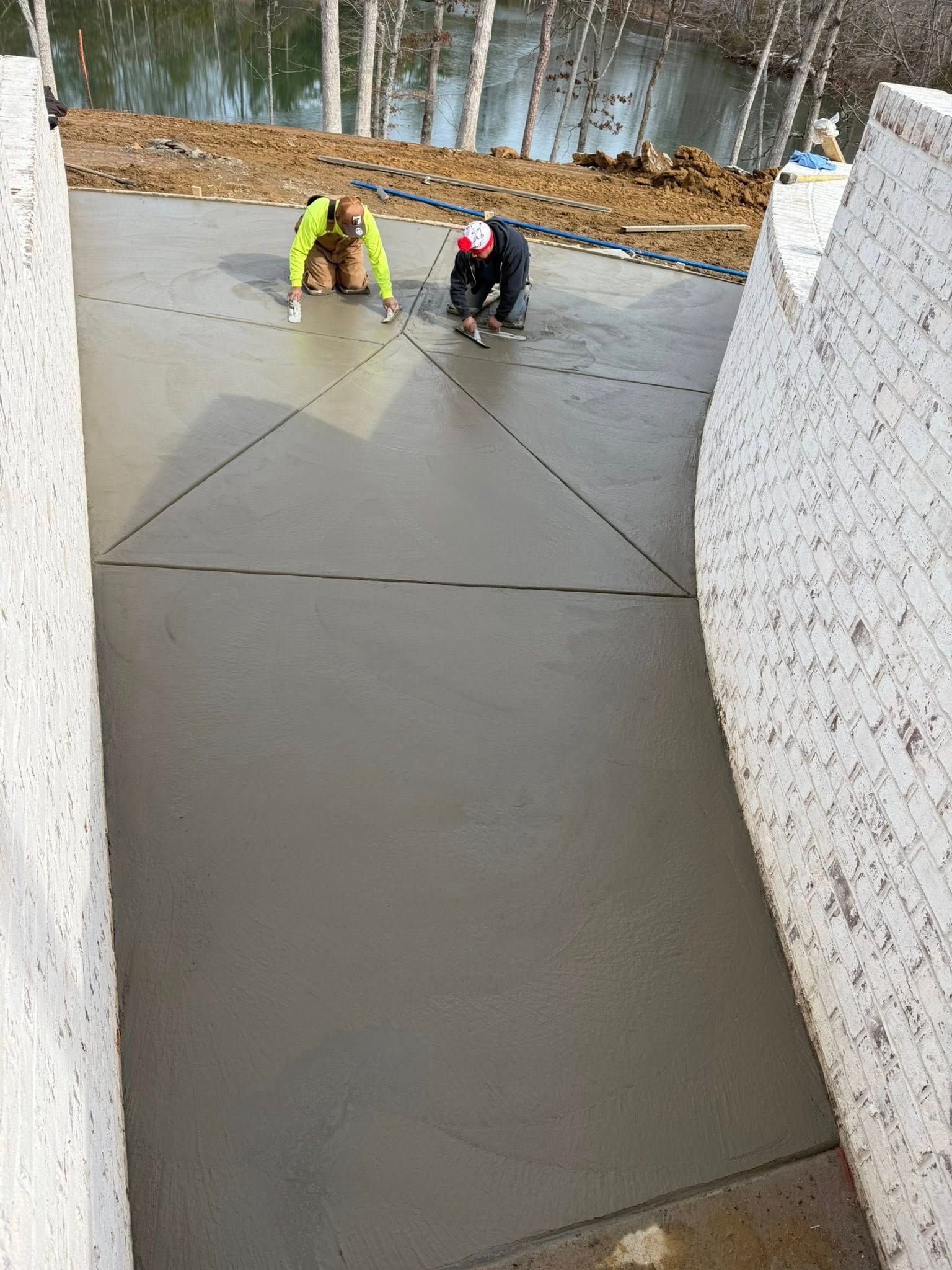 Workers smoothing wet concrete on a slab outdoors beside a stone wall and woods.