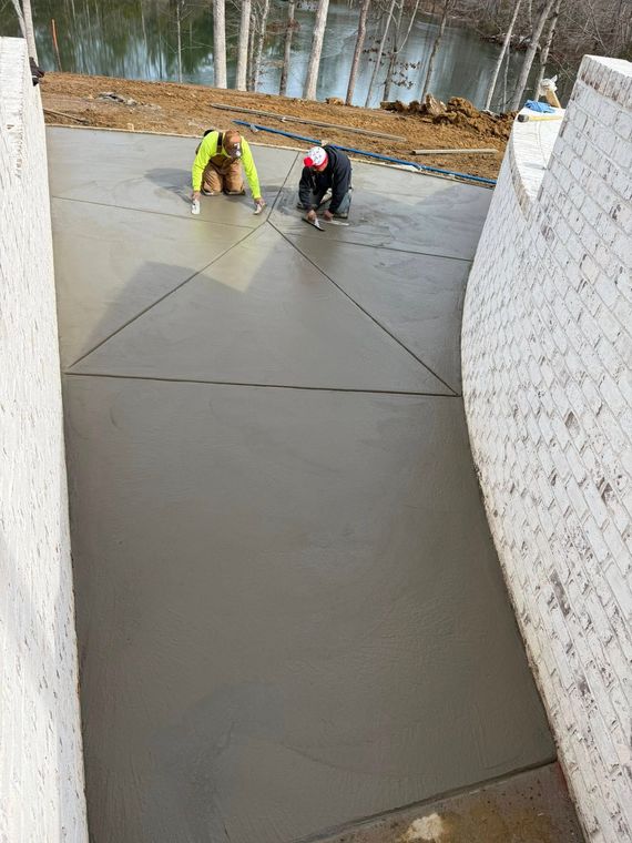 Workers smoothing wet concrete on a slab outdoors beside a stone wall and woods.