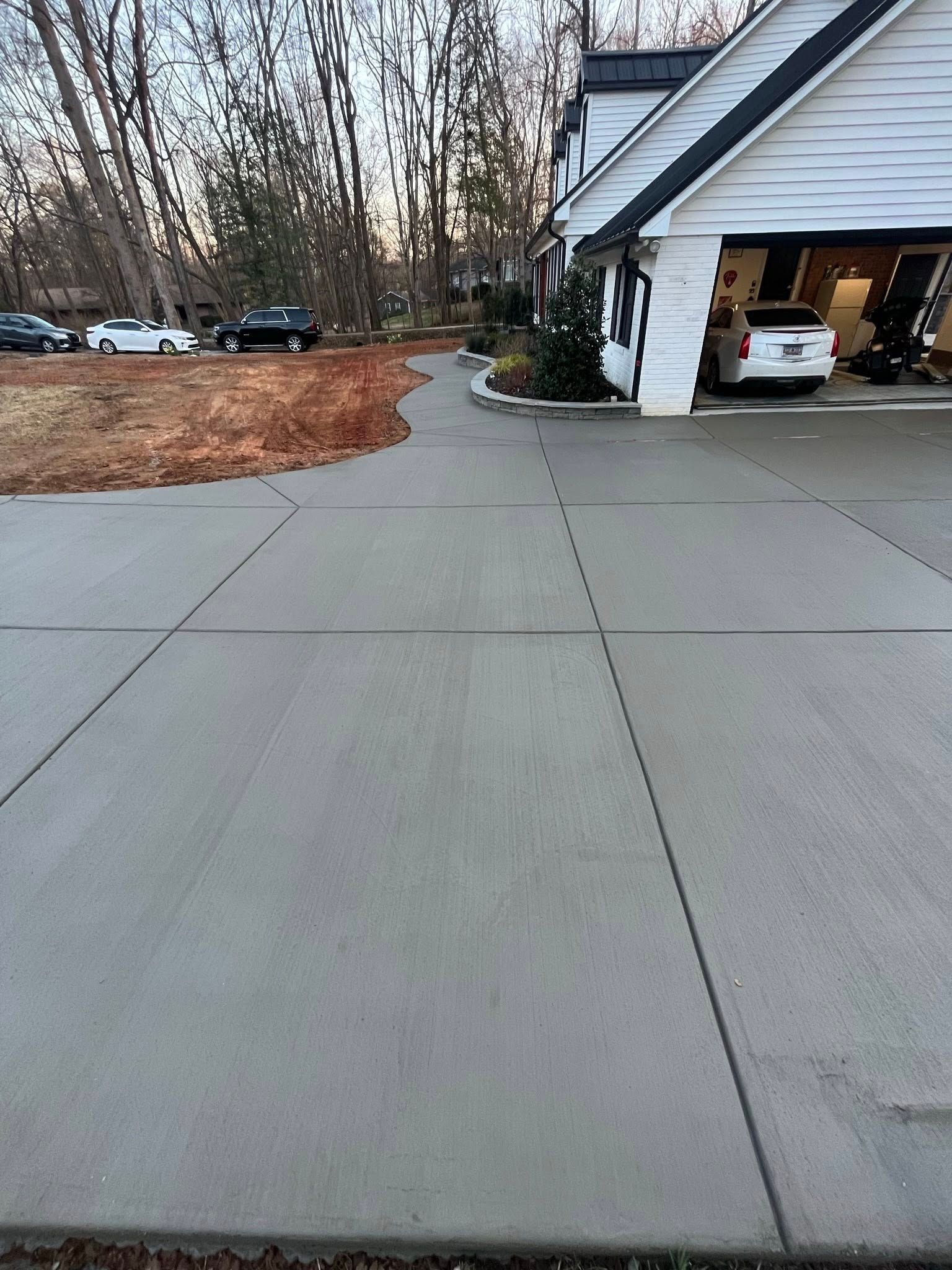 Wide concrete driveway leading to an open garage beside a white house, with a wooded yard in winter.