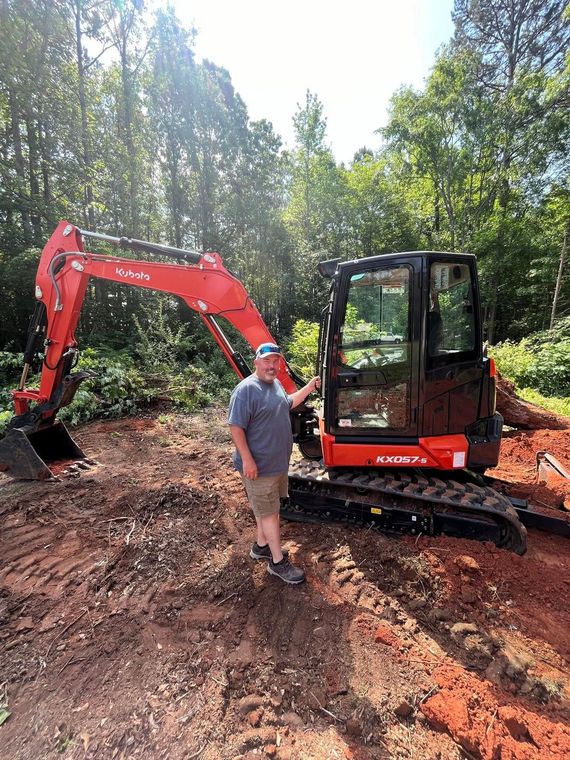 Red excavator with person standing beside it on a dirt lot surrounded by trees.
