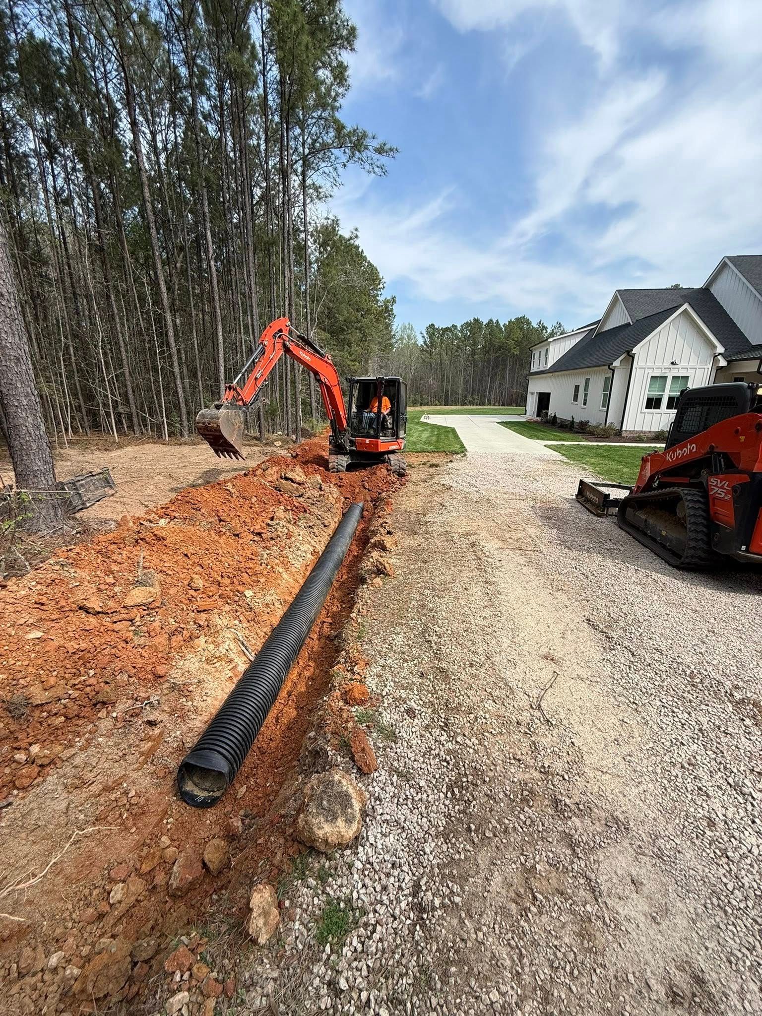 Construction crew laying black drainage pipe along a gravel driveway beside a house and trees